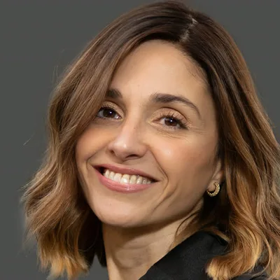 Lital Shahar smiling, shoulder-length brown hair, in a black top against a grey background